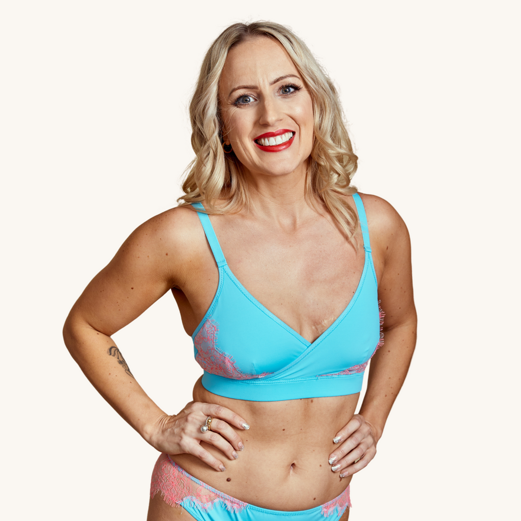 A smiling blond woman wears our Icon blue cross over bralette featuring blush pink lace - Valiant Lingerie