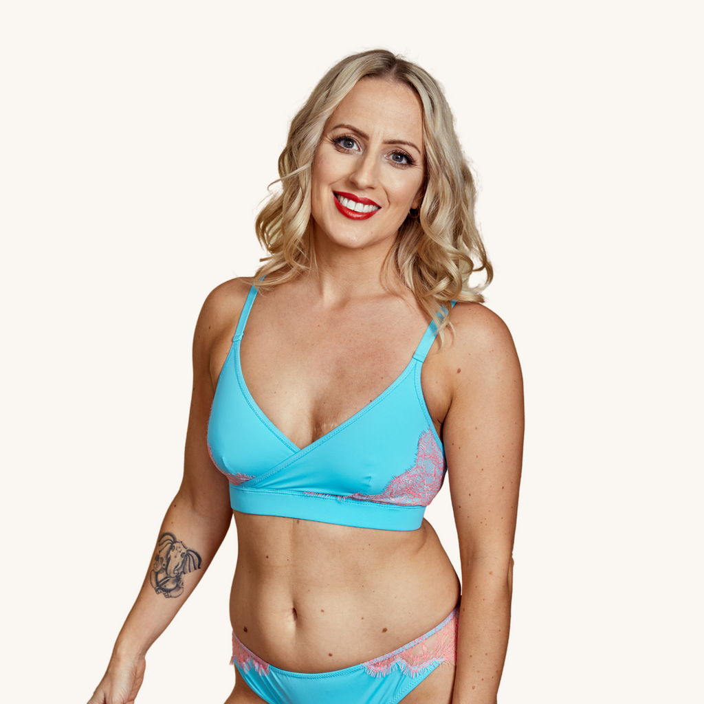 A smiling blond woman wears our Icon blue cross over bralette with blush pink lace- Valiant Lingerie